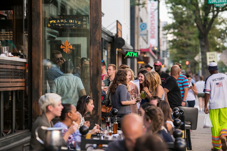 A lively crowd gathers outside Pearl Dive Oyster Palace, mingling and enjoying drinks along a bustling 14th Street sidewalk.